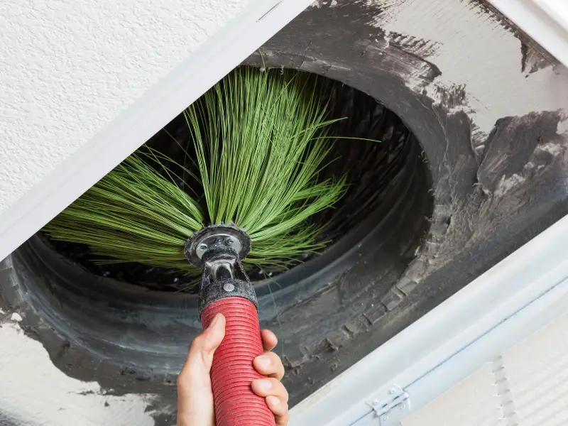 A hand holds a red rotary brush with green bristles inside a dusty HVAC air duct during professional air duct cleaning.