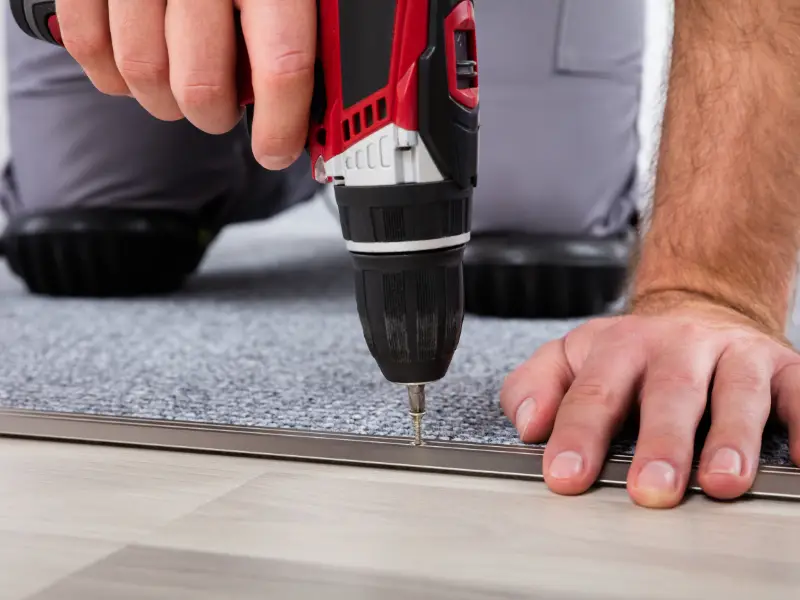 A technician uses a red drill to fasten trim at the edge of a gray carpet, with one hand steadying the floor during carpet restretching.