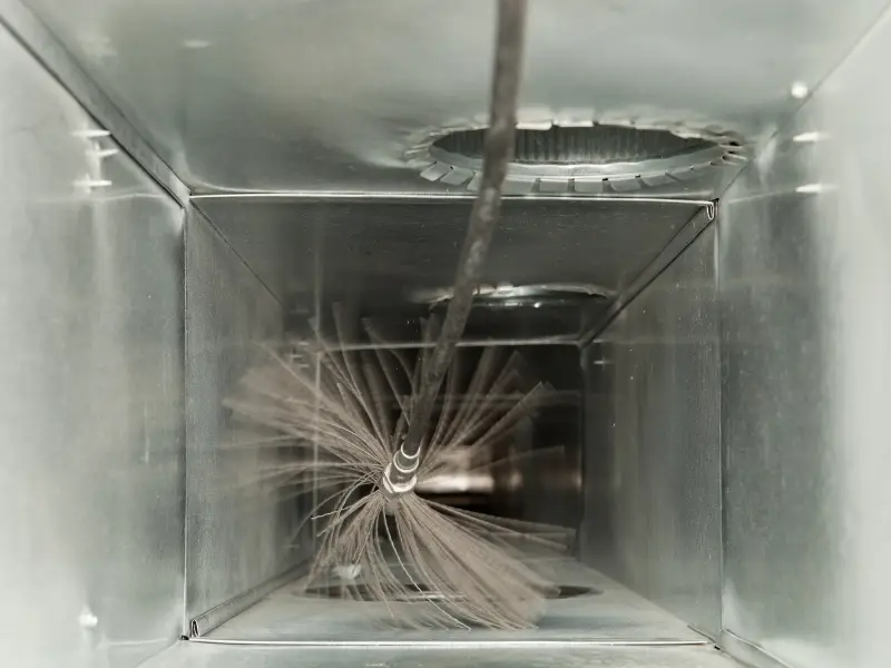 A rotary brush sweeps through a metal HVAC duct, clearing built-up dust and debris from the interior walls during air duct cleaning.