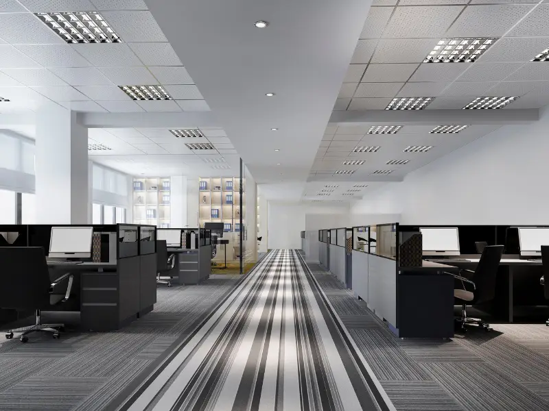 A bright commercial office shows rows of workstations, glass partitions, and a striped carpet corridor beneath a grid of ceiling lights.