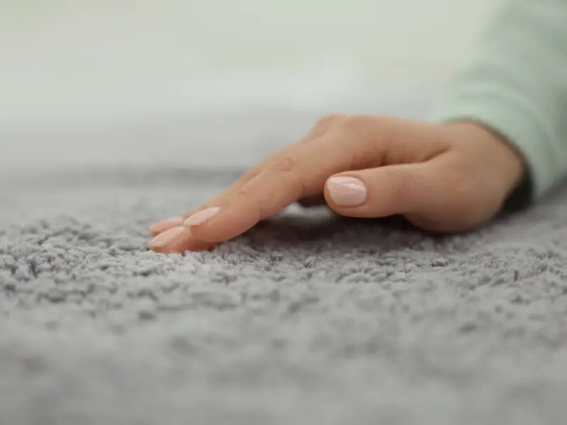 A hand gently presses into a gray carpet, showing the soft pile and clean, well-kept texture in a quiet indoor setting with a fresh appearance.