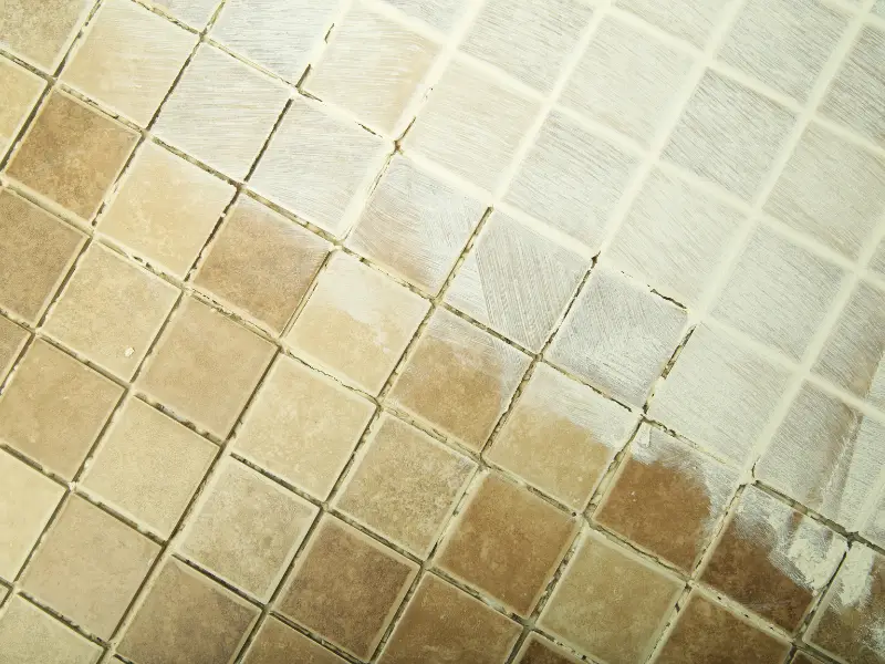 Half-cleaned beige tile floor shows dirty grout lines beside a bright white cleaned section after professional tile cleaning work.