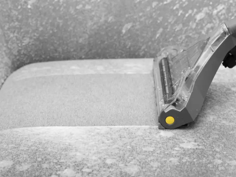 A gray upholstery surface is being cleaned with an extraction nozzle, lifting dirt from the fabric on a sofa and leaving a fresh path behind.